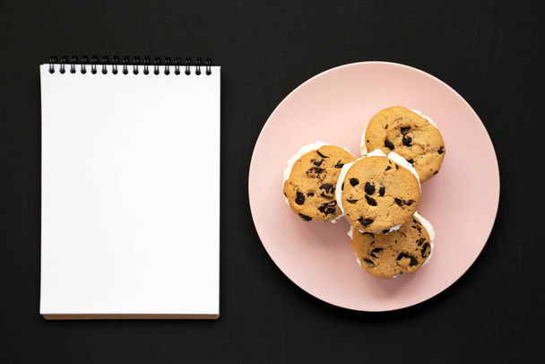 Homemade Chocolate Chip Cookie Ice Cream Sandwich on a pink plate, blank notepad on a black background, top view. Overhead, from above, flat lay. - Foto, immagini