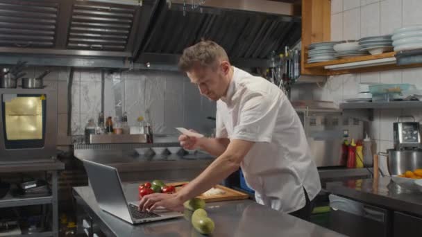 Hidasliikkeinen keskipitkän kuvamateriaalia valkoihoinen kokki kaupallisessa keittiössä kannettavan tietokoneen samalla ruoanlaitto terve ruokalaji tilaus lippu - Materiaali, video