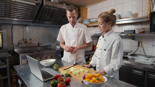 Medium shot of skilled chef teaching young female cook basics of cutting with chef knife, showing video tutorial on laptop during practice at restaurant kitchen - Footage, Video