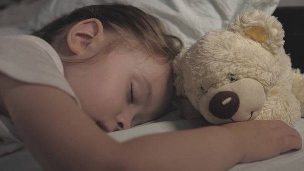 Το παιδί κοιμάται στο σπίτι στον καναπέ στο παιδικό δωμάτιο. Το μωρό που κοιμάται είναι χαρούμενο και ανέμελο στο κρεβάτι, αγκαλιάζοντας ένα αρκουδάκι. Η μητέρα κάλυψε το παιδί της με μια κουβέρτα. Ευτυχία σε ένα όνειρο. - Φωτογραφία, εικόνα