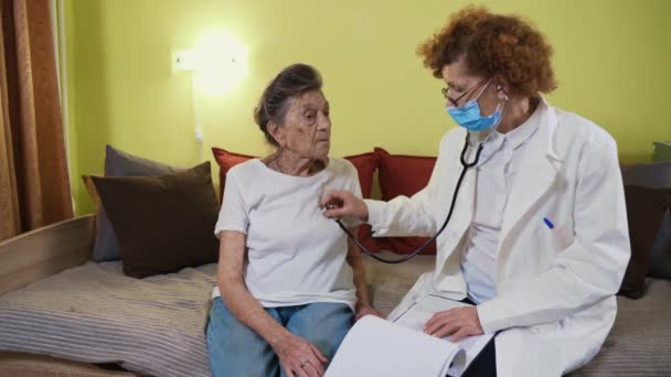 Beyaz önlüklü yaşlı kadın doktor, tıbbi maske karantina ve koronavirüs sırasında yaşlı kadını muayene ediyor. Sıhhiye akciğer ve kalp hastalarını kontrol etmek için steteskop kullanıyor. - Video, Çekim
