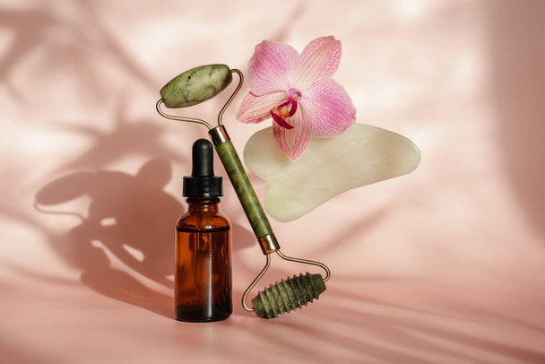 Photo for the spa salon. Roller and Guasha scraper for facial massage, massage oil, black hot spa stones, pink orchids, white towel on a pink background. Balance and levitation trending style - Photo, Image