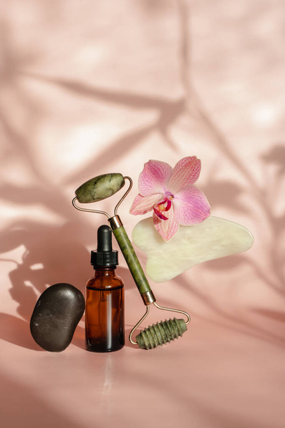 Photo for the spa salon. Roller and Guasha scraper for facial massage, massage oil, black hot spa stones, pink orchids, white towel on a pink background. Balance and levitation trending style - Photo, Image