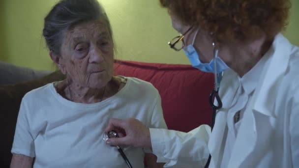 Femme âgée médecin en blouse blanche, masque médical fait l'examen médical de la femme âgée à la maison pendant la quarantaine et le coronavirus. Le médecin utilise un stéthoscope pour vérifier les poumons et le cœur d'un patient âgé - Séquence, vidéo