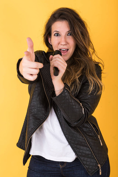 Young singer girl, wearing black leather jacket, singing with microphone and pointing with her hand on yellow background. - Photo, Image