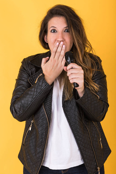 Νεαρή τραγουδίστρια, φορώντας μαύρο δερμάτινο μπουφάν και μικρόφωνο, κάνοντας χειρονομία έκπληξη στο κίτρινο φόντο. - Φωτογραφία, εικόνα