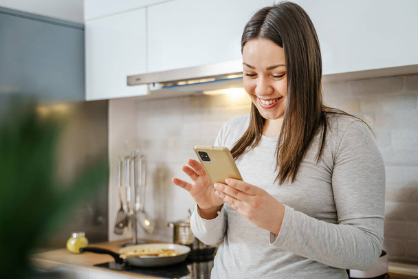 Portret młodej białej kobiety w kuchni przy użyciu telefonu komórkowego przeglądającej internet dla przepisu online podczas gotowania posiłku lub rozmowy wideo - przestrzeń do kopiowania - Zdjęcie, obraz