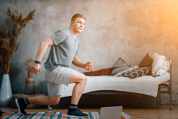 Молодой человек занимается спортом дома, онлайн тренировкой с ноутбука. Спортсмен выпрыгивает, смотрит кино в спальне, на заднем плане кровать, ваза, ковер. - Фото, изображение