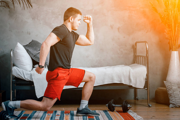 A young man goes in for sports at home, online workout from the laptop. The athlete lunges, watches a movie  in the bedroom, in the background there is a bed, a vase, a carpet. - Foto, Imagem