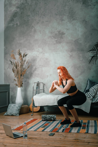 The beauty woman goes in for sports at home. Cheerful sporty woman with red hair  doing a squat   and watches  in laptop, shootting  blog  in the bedroom - Photo, image