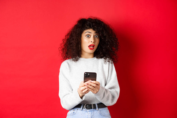 Image of pretty curly woman with shocked face, reading message on smartphone startled, standing against red background - Valokuva, kuva