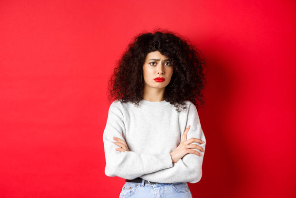 Sad and worried caucasian woman frowning, cross arms on chest and looking concerned, feeling bad, standing on red background - Foto, Imagen
