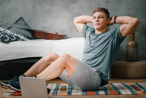 Young man goes in for sports at home, training online. The athlete makes the press, smiles, there is an open laptop, bed, vase, carpet nearby. - Foto, imagen