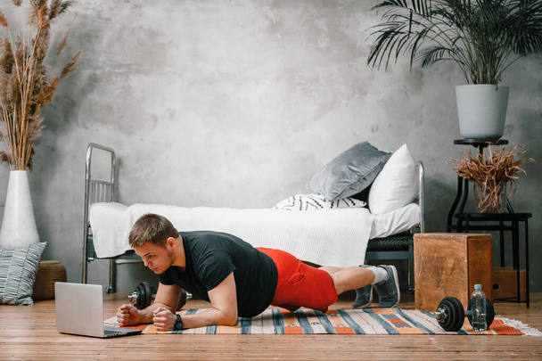 Young man goes in for sports at home, training online. The athlete makes a plank, watches a movie and  social networks on laptop  in the bedroom, in the background a bed, a vase, a carpet. - Photo, Image