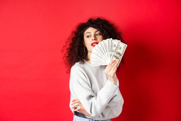 Bella donna ricca guardando sensuale da parte, agitando a se stessa con dollaro banconote ventilatore, in piedi seducente su sfondo rosso - Foto, immagini