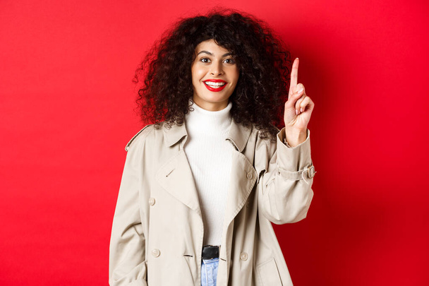 Belle femme moderne avec des lèvres rouges, coiffure bouclée, portant un trench-coat de printemps, doigt pointant vers le haut et souriant, debout sur fond rouge - Photo, image