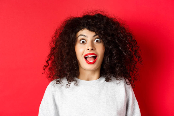 Close-up portrait of excited woman with curly hair, scream surprised and amazed, checking out special deal, standing on red background - Photo, image