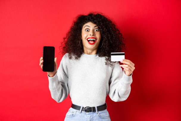 Achats en ligne. Joyeux jeune femme montrant carte de crédit en plastique et écran de téléphone vide, annoncer l'offre promo, debout sur fond rouge - Photo, image