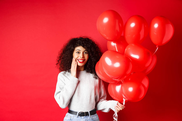 Holidays and celebration. Surprised birthday girl holding helium party balloons and looking touched at camera, being congratulated, red background - Foto, Bild
