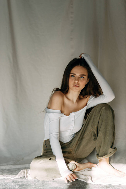 Model tests of a pretty girl in basic clothing on fabric. Portrait of a young skinny brunette posing in a white bodysuit, sitting on a white floor in a white studio with shadows from the window. - Foto, Imagen