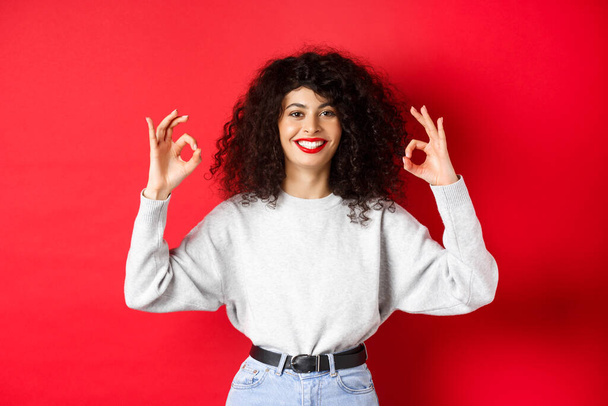 Confident pretty girl with curly hairstyle, showing okay gestures and smiling, approve and agree with you, praising excellent choice, standing satisfied on red background - Photo, Image