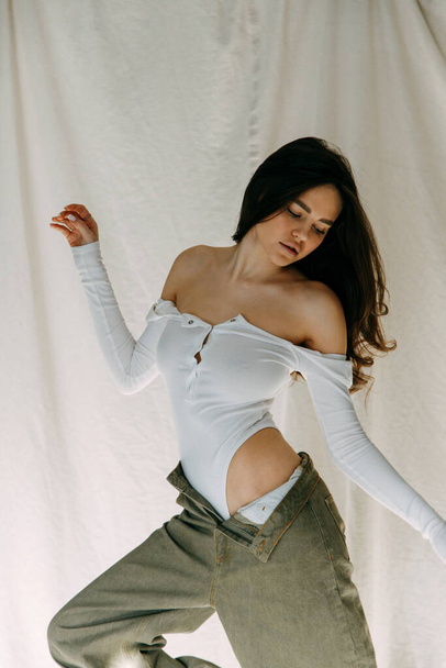 Model tests of a pretty girl in basic clothing on fabric. Portrait of a young skinny brunette posing in a white bodysuit, sitting on a white floor in a white studio with shadows from the window. - Фото, изображение