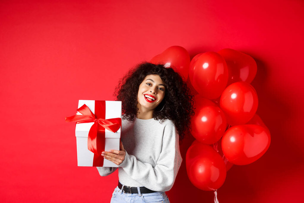Праздники и праздники. Счастливая именинница, держа в руках подарок и позируя возле праздничных шаров, возбужденно улыбаясь на камеру, на красном фоне - Фото, изображение