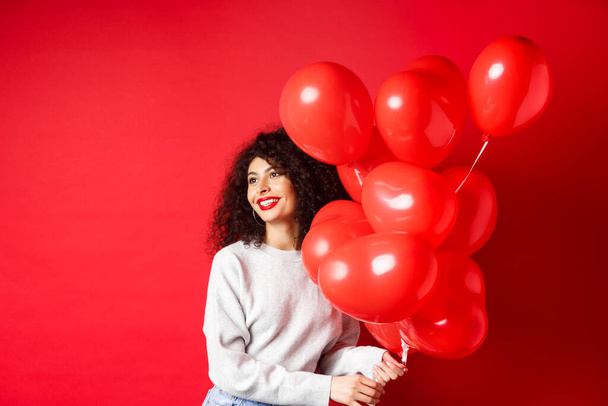Свята та святкування. Щаслива жінка позує з вечірніми кульками на червоному тлі, дивлячись убік на порожнє місце
 - Фото, зображення