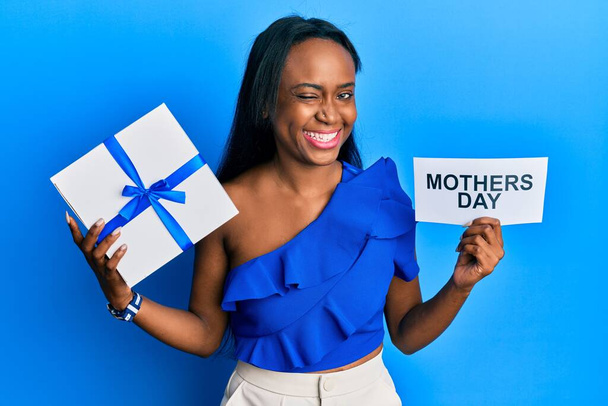 Young african woman holding gift and mothers day text winking looking at the camera with sexy expression, cheerful and happy face.  - Fotografie, Obrázek