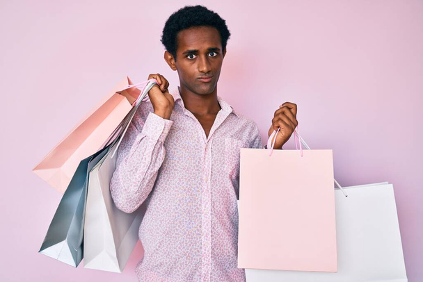 Hombre guapo africano sosteniendo bolsas de compras escéptico y nervioso, frunciendo el ceño molesto debido a un problema. persona negativa.  - Foto, imagen