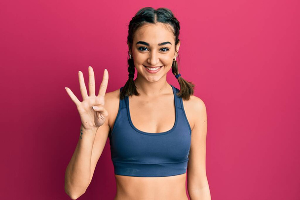 Młoda brunetka dziewczyna w odzieży sportowej i warkocze pokazując i wskazując palcami numer cztery, uśmiechając się pewny siebie i szczęśliwy.  - Zdjęcie, obraz