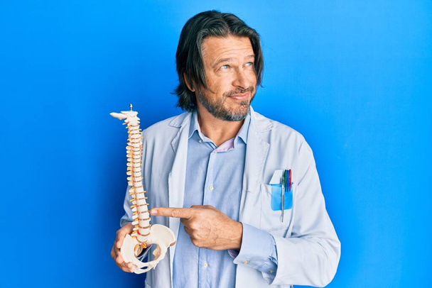 Középkorú jóképű traumatológus férfi kezében anatómiai modell gerincoszlop mosolyog oldalra néz, és bámul félre gondolkodás.  - Fotó, kép