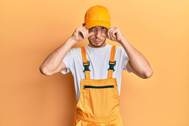 Hispanic young man wearing handyman uniform trying to open eyes with fingers, sleepy and tired for morning fatigue  - Photo, Image