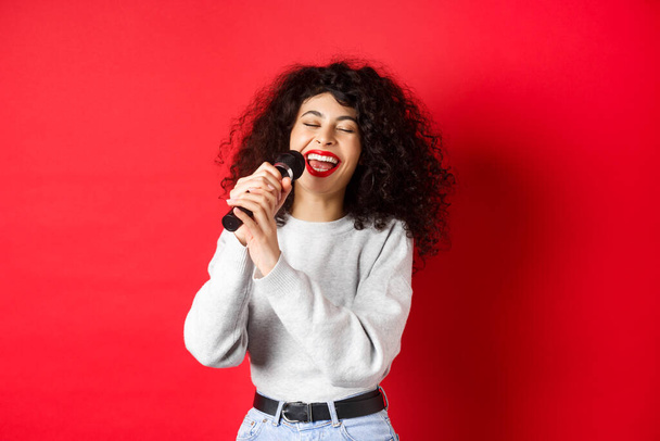 Zainteresowania i wypoczynek. Szczęśliwa kobieta śpiewająca piosenkę w mikrofonie, bawiąca się na karaoke z mikrofonem, stojąca na czerwonym tle - Zdjęcie, obraz