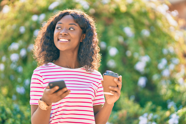 Chica afroamericana joven usando teléfono inteligente beber café en el parque. - Foto, Imagen