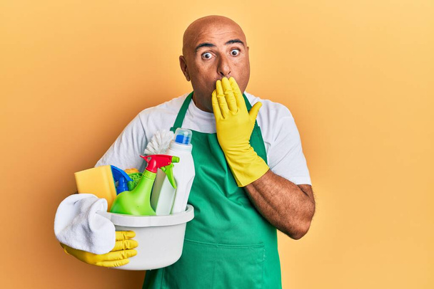 Mature middle east man wearing cleaner apron holding cleaning products covering mouth with hand, shocked and afraid for mistake. surprised expression  - Fotó, kép