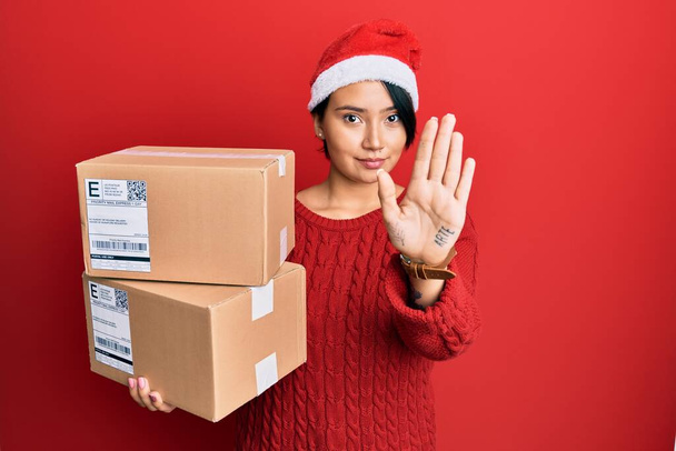 Beautiful young woman with short hair wearing christmas hat holding delivery packages with open hand doing stop sign with serious and confident expression, defense gesture  - Foto, afbeelding