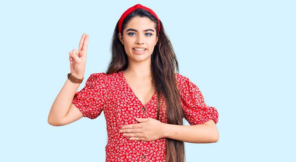 Brunette teenager girl wearing summer dress smiling swearing with hand on chest and fingers up, making a loyalty promise oath  - Φωτογραφία, εικόνα