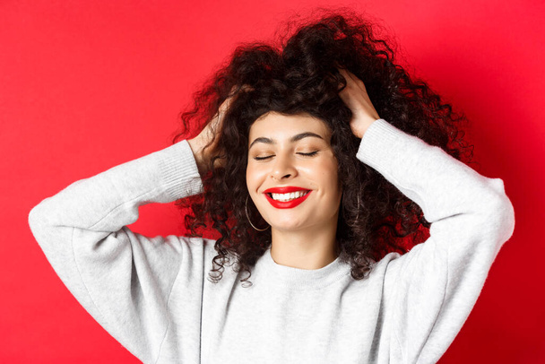Κοντινό πλάνο της ανέμελης γυναίκας αγγίζοντας μαλακά σγουρά μαλλιά και χαμογελώντας ευχαριστημένος, στέκεται σε κόκκινο φόντο. έννοια της περιποίησης και της ομορφιάς - Φωτογραφία, εικόνα