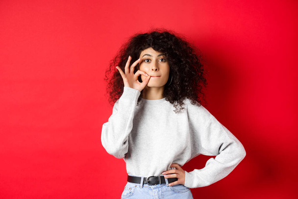 Image of cute girl with curly hair making promise to stay quiet, zipping lips, making seal, hiding a secret, standing dumb on red background - Fotoğraf, Görsel