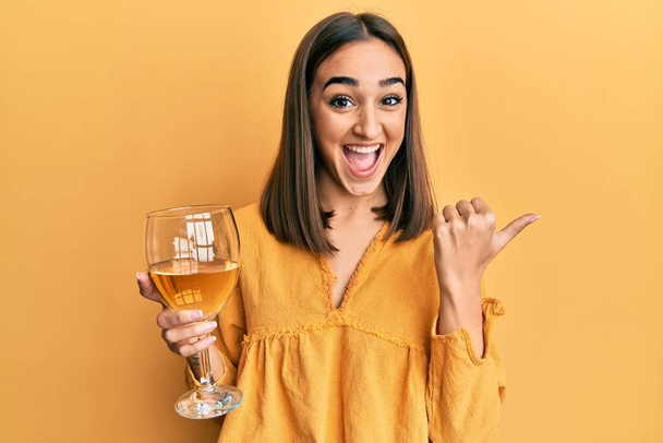 Молода брюнетка п'є склянку білого вина вказуючи великий палець в сторону усміхнений щасливий з відкритим ротом
  - Фото, зображення