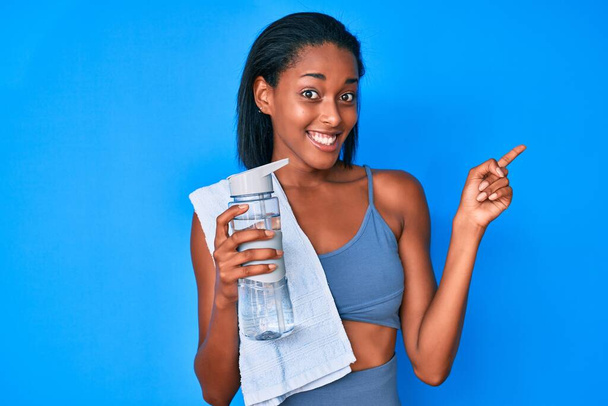 Jeune femme afro-américaine portant des vêtements de sport boire bouteille d'eau souriant heureux pointant avec la main et le doigt sur le côté  - Photo, image
