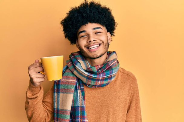 Young african american man with afro hair drinking a cup of coffee looking positive and happy standing and smiling with a confident smile showing teeth  - Фото, зображення