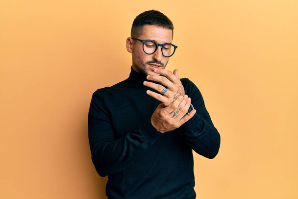 Όμορφος άντρας με τατουάζ που φοράει ζιβάγκο πουλόβερ και γυαλιά που υποφέρουν από πόνο στα χέρια και τα δάχτυλα, φλεγμονή αρθρίτιδα  - Φωτογραφία, εικόνα