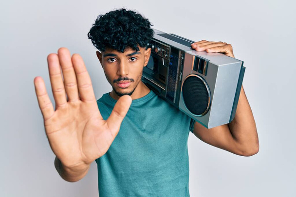 Jonge Arabische knappe man met boombox, luisteren naar muziek met open hand doen stopbord met serieuze en zelfverzekerde uitdrukking, verdediging gebaar  - Foto, afbeelding