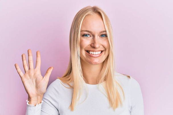 Junges blondes Mädchen in lässiger Kleidung zeigt mit Finger Nummer fünf nach oben und lächelt dabei selbstbewusst und glücklich.  - Foto, Bild