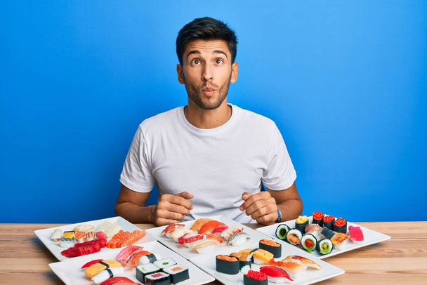 Młody przystojniak jedzący sushi na stole, robiący rybią twarz ustami, szalony i komiczny gest. śmieszne wyrażenie.  - Zdjęcie, obraz