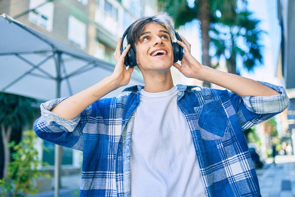 Νεαρός Ισπανός χαμογελάει χαρούμενος με ακουστικά που περπατάνε στην πόλη. - Φωτογραφία, εικόνα