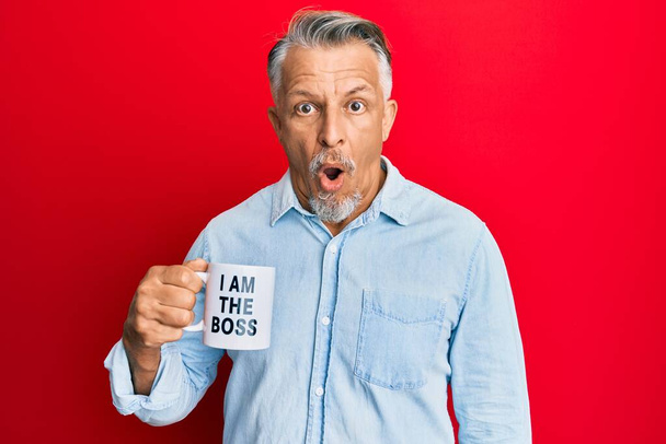Седовласый мужчина средних лет, пьющий из я босс кофе чашку испуганный и пораженный с открытым ртом для удивления, неверие лицо  - Фото, изображение