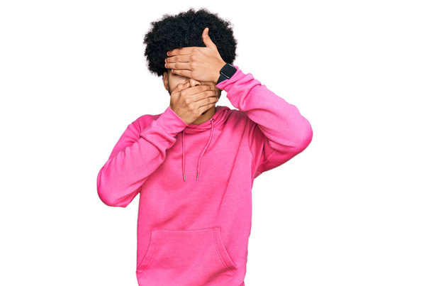 Молодой африканский американец с африканскими волосами, одетый в обычную розовую толстовку, прикрывающую глаза и рот руками, удивлен и шокирован. скрывая эмоции  - Фото, изображение